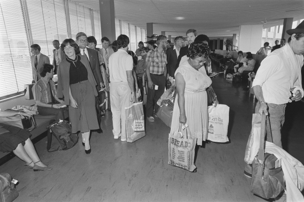 Schwarzes und weißes Bild von Reisenden in einem Flughafen, einige gehen mit Gepäck, andere sitzen auf Bänken oder stehen, mit einem Mann und einer Frau, die Taschen halten, in der Mitte und Fensterläden auf der linken Seite.