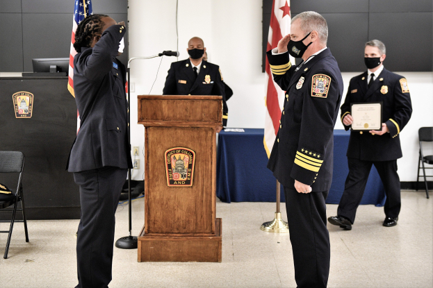 Eine Gruppe uniformierter Männer steht vor einem Podium mit einem Mikrofon, flankiert von einer sitzenden Person links und zwei stehenden Individuen rechts, einer haltend ein Zertifikat, während einer Feuerwehrabendverleihung mit Flaggen, einem Tisch mit einer blauen Decke, einem Monitor und anderen Gegenständen im Hintergrund.