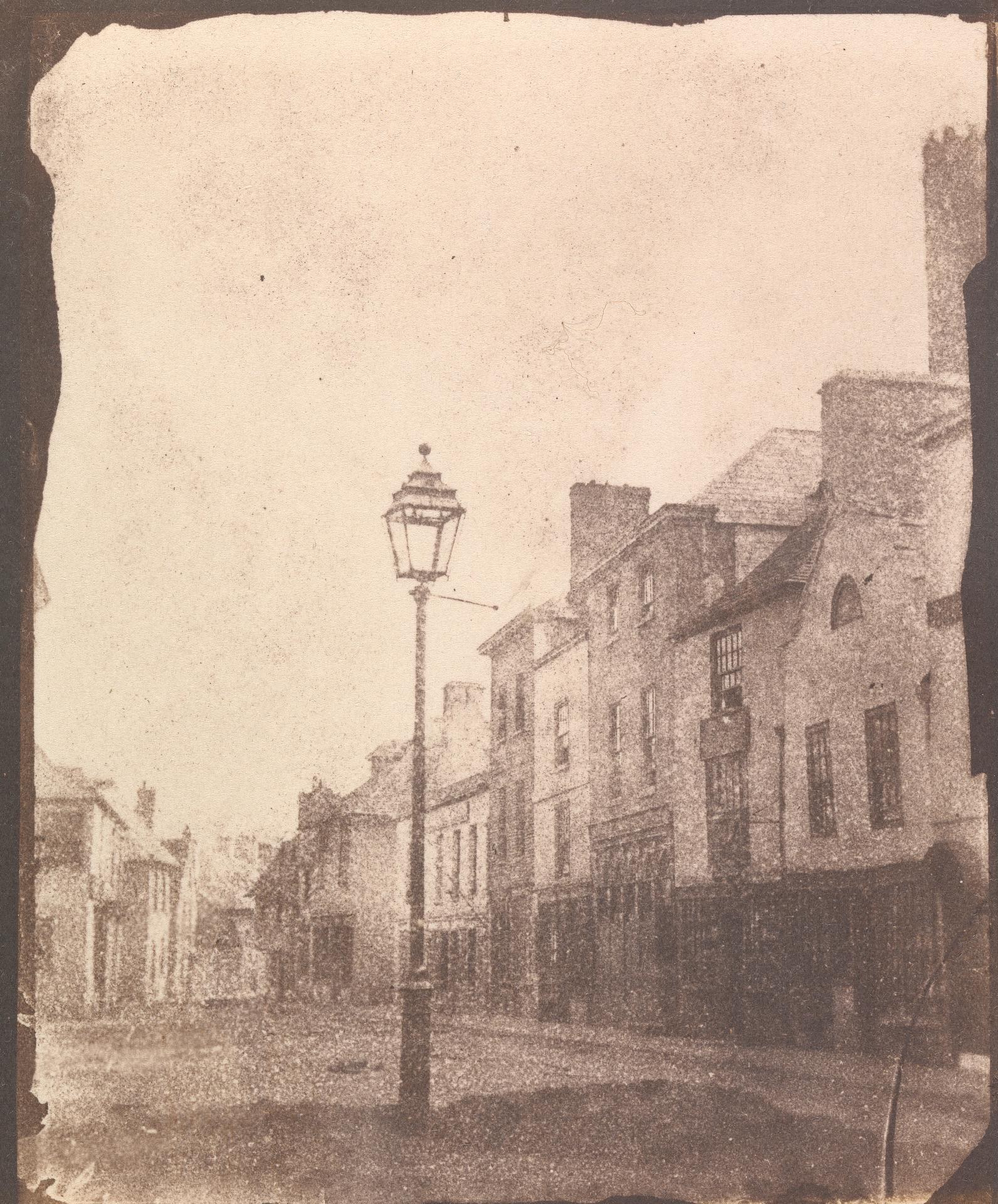 Alte Schwarz-Weiß-Fotografie einer Straßenlaterne an einer Kreuzung, flankiert von Gebäuden unter einem klaren Himmel.