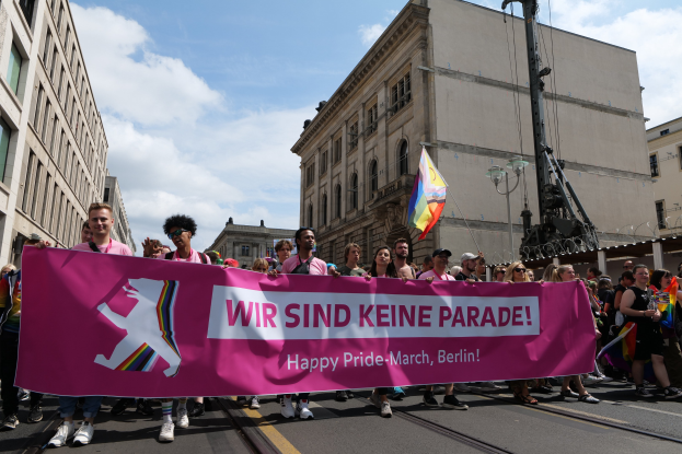 Gruppe von Menschen, die eine Straße in Berlin, Deutschland, entlanggehen und ein rosanes Banner mit der Aufschrift "Happy Pride March" halten, mit Gebäuden auf beiden Seiten und einem Fahnenmast im Vordergrund unter einem bewölkten Himmel.