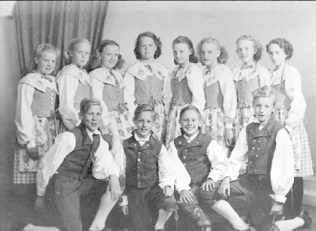 Schwarzes und weißes Foto von Kindern, die vor einem Vorhang posieren, mit dem Text "1930er Kinder-Schule" unten.