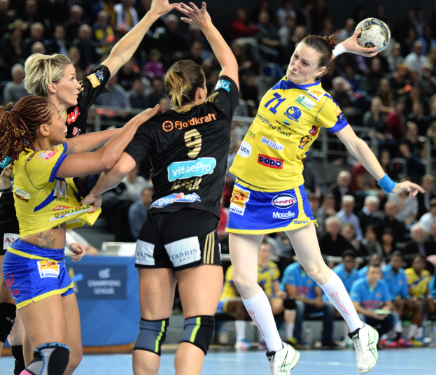 Eine Gruppe von Frauen beim Handballspielen, eine hält den Ball, mit unscharfen Zuschauern und Text im Hintergrund.