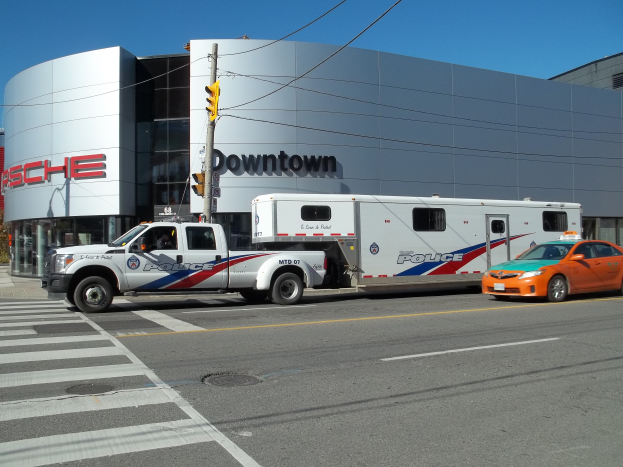 Ein Polizeilaster steht vor einem Gebäude mit Text darauf, mit einer Verkehrsampel mit einem Mast und Drähten im Vordergrund und Bäumen und einem klaren blauen Himmel im Hintergrund.
