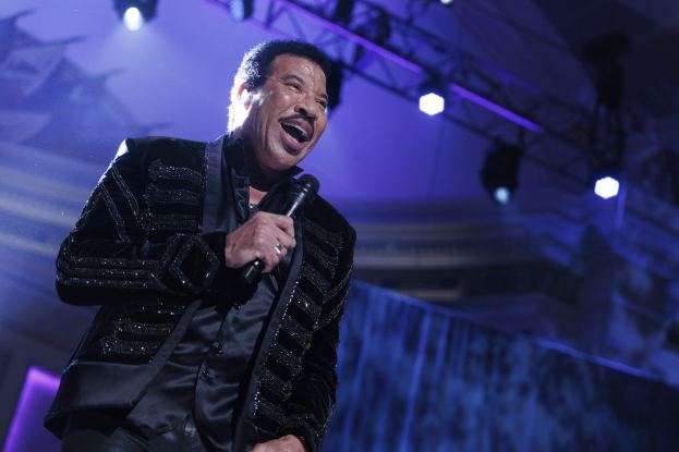 Lionel Richie performs bei den Grammys in einem schwarzen Kleid und hält ein Mikrofon in der Hand, während helle Bühnenlichter eine dramatische Atmosphäre schaffen.