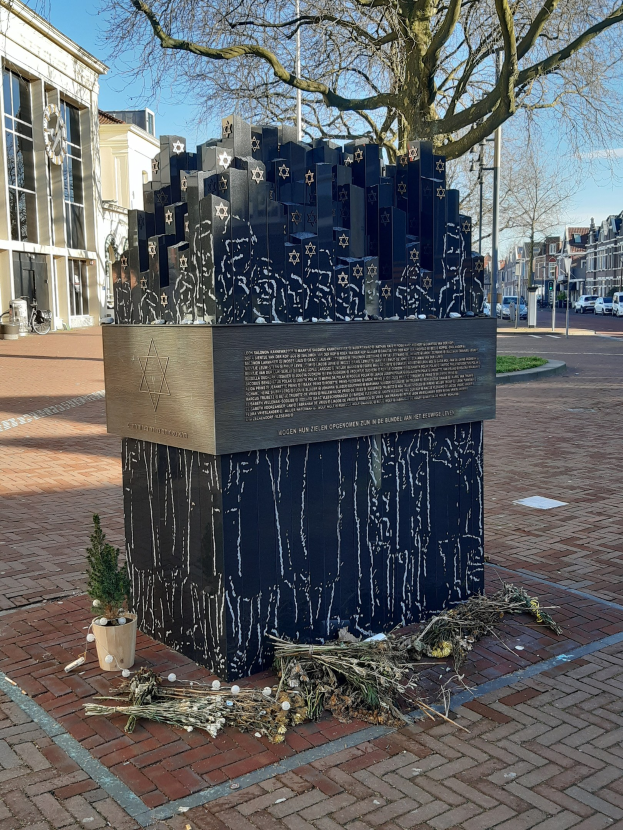 Denkmal für Holocaust-Opfer in einem Stadtplatz mit einem Baum davor, einer Topfpflanze, verstreuten trockenen Blättern und umliegenden Gebäuden, Fahrzeugen, Polen und Bäumen.