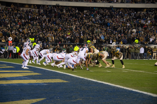 Ein Fuballspiel im Gange auf einem Stadionfeld mit Spielern in Sportbekleidung und Helmen, umgeben von Zuschauern und Stadionelementen.