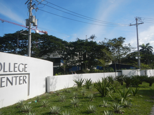College Center-Eingangsschild an einer Wand umgeben von Grün, Weg, Strommasten, Bäumen, Gebäuden, einem Kran und einem bewölkten Himmel.