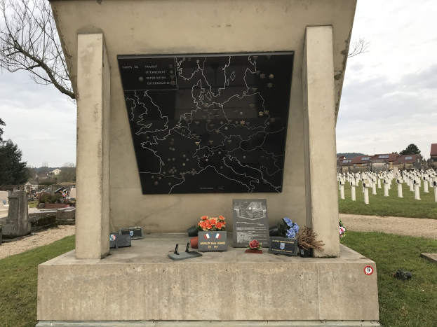 Grabmal eines deutschen Soldaten in einem Friedhof mit einer Karte, umgeben von Blumen und Gegenständen, mit Gräbern, Bäumen, Gebäuden und einem blauen Himmel im Hintergrund.