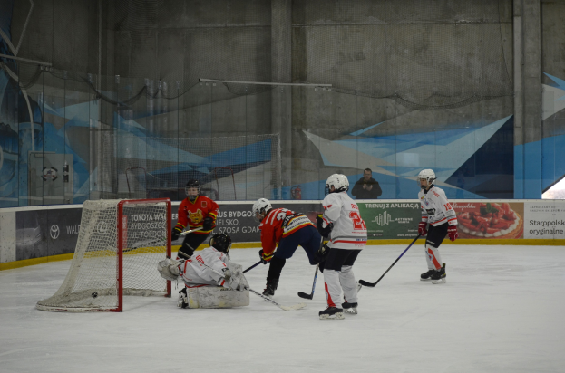 Gruppe von Menschen, die Eisstockschießen auf einem Eisstadion mit Helmen, Eishockeyschlägern und einem Tor auf der linken Seite, mit Bannern und einer Wand im Hintergrund.