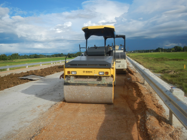 Straßenwalze legt Asphalt auf der Seite einer Straße mit Geländer, Gras, Bäumen und bewölktem Himmel im Hintergrund.