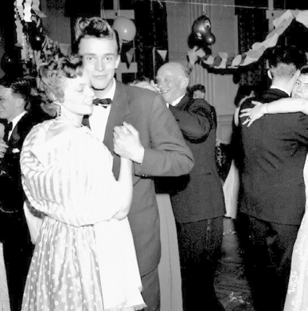 Ein Schwarz-Weiß-Foto eines Mannes und einer Frau, die auf einer Party tanzen, umgeben von lachenden Zuschauern und Ballons sowie Dekorationen im Hintergrund.