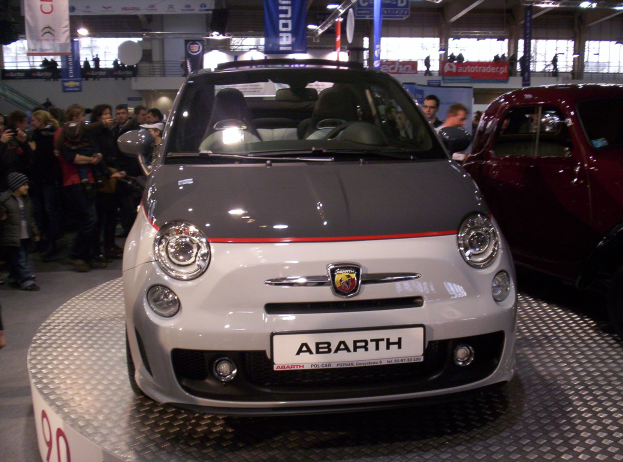 Ein roter und schwarzer Fiat 500 Abarth auf einer Autauschstellung umgeben von Menschen mit Bannern und Beleuchtung im Hintergrund.