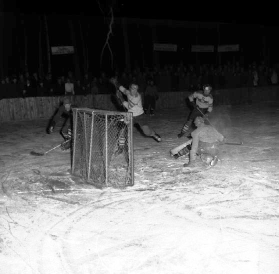 Schwarzes und weißes Bild eines Hockey-Spiels auf einem Eisstadion, mit Spielern in der Nähe eines Tores und Zuschauern im Hintergrund.