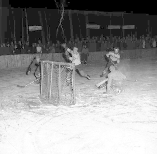 Schwarzes und weißes Bild eines Hockey-Spiels auf einem Eisstadion, mit Spielern in der Nähe eines Tores und Zuschauern im Hintergrund.