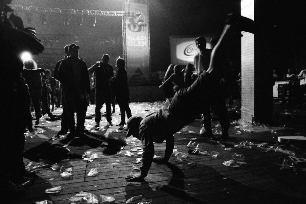 Eine Person, die in der Mitte eine Stunt-Nummer ausführt, umgeben von anderen Personen, Plastikabfällen auf dem Boden, einer Tafel mit Text und Scheinwerfern von oben.