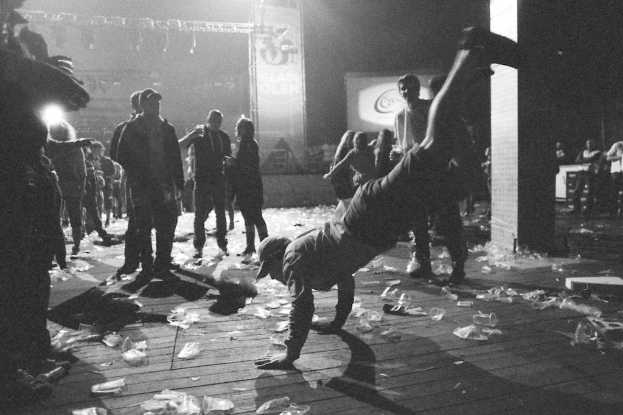 Eine Person, die in der Mitte eine Stunt-Nummer ausführt, umgeben von anderen Personen, Plastikabfällen auf dem Boden, einer Tafel mit Text und Scheinwerfern von oben.