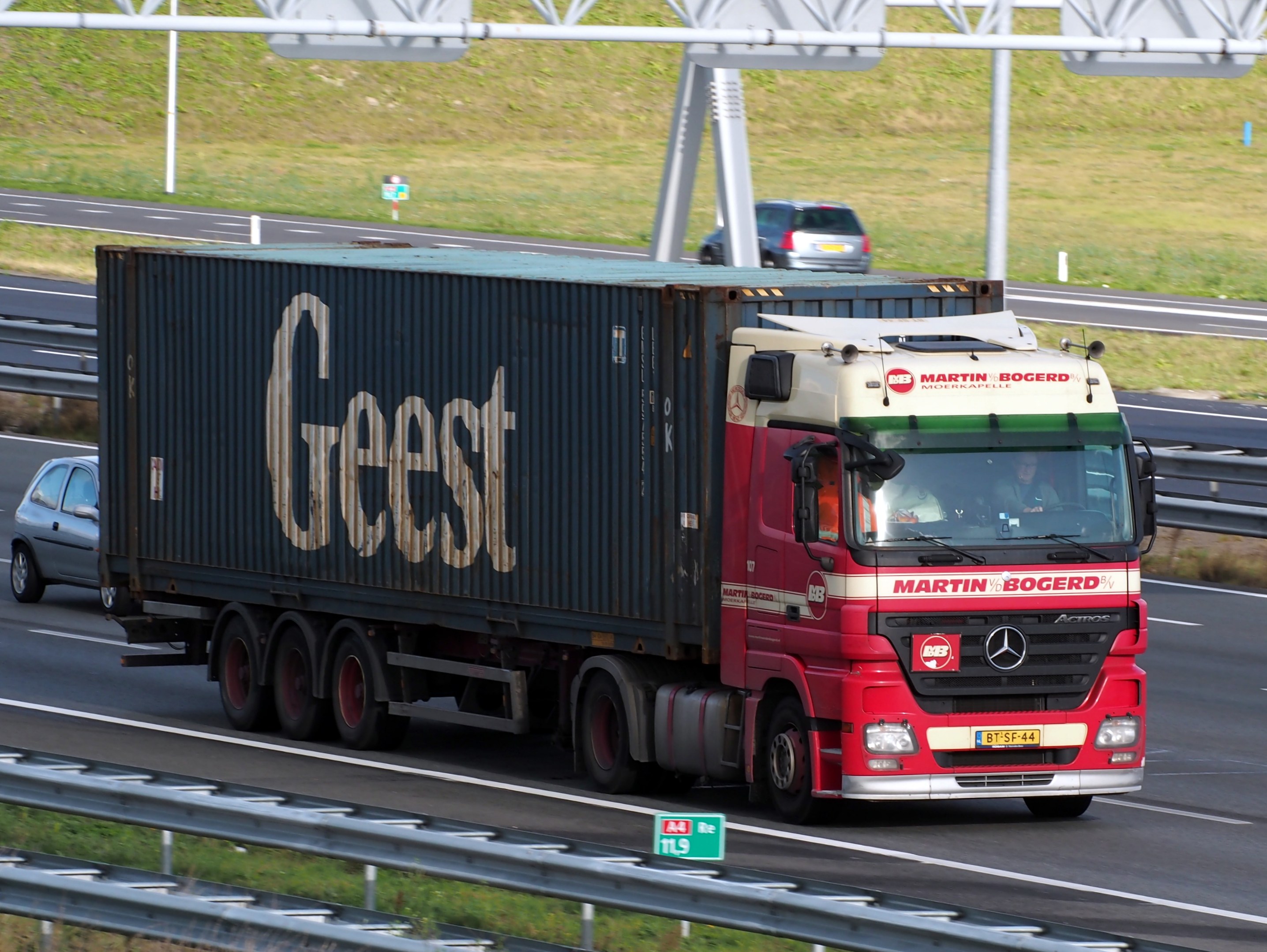 Roter Lastwagen auf einer Autobahn mit einem Container auf dem Rücken, mehrere Personen im Inneren und Strommasten, Holzplanken und Gras im Hintergrund.