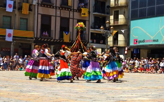 Eine Gruppe von Frauen in bunten Kleidern, die auf der Straße tanzen und von einer Menschenmenge umgeben sind, mit Gebäuden, Geländern, Balkonen, Fahnen, einem Laternenpfahl und einer Tafel mit Schrift im Hintergrund.