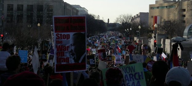 Menschen protestieren auf einer Straße und halten Schilder, mit Gebäuden, Bäumen, Laternenmasten und dem Himmel im Hintergrund.