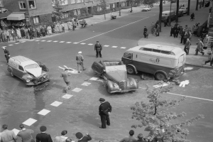Eine Schwarz-Weiß-Szene eines Autounfalls an einer Straßenkreuzung mit mehreren Fahrzeugen einschließlich eines Lieferwagens, einer Gruppe von Menschen drumherum und Gebäuden, Bäumen, Pfählen und Texttafeln im Hintergrund.