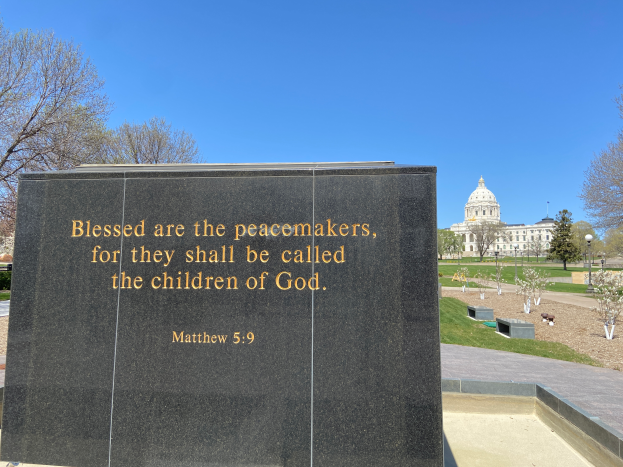 Ein Gedenkstein mit der Inschrift "Selig sind die Friedfertigen, denn sie sollen das Reich Gottes erben" umgeben von Bäumen, Pflanzen, Gras, Laternenmasten und einem Gebäude unter einem klaren blauen Himmel.