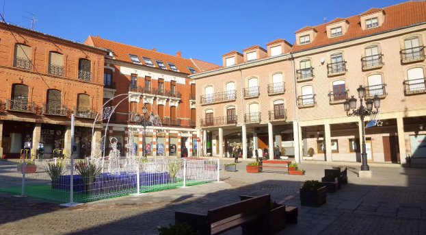 Ein Stadtplatz mit einem zentralen Brunnen, umgeben von Bänken, Topfpflanzen, einem Metallzaun, Straßenlaternen, Menschen, Schildern, Gebäuden mit Fenstern unter einem bewölkten Himmel.