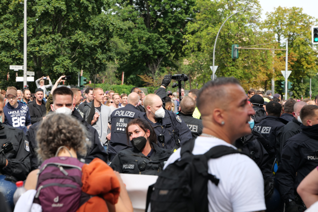 Eine große Gruppe von Menschen steht vor einer Reihe von Polizeibeamten, einige tragen Masken und Rucksäcke, andere halten Kameras, mit Laternenmasten, Verkehrssignalen, Schildern, Bäumen und einem klaren blauen Himmel im Hintergrund.
