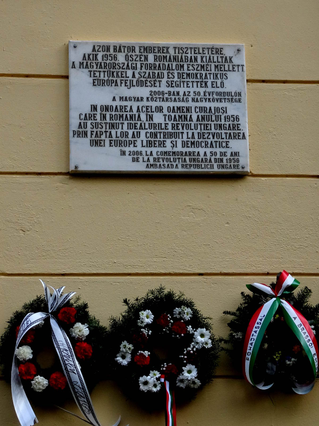 Zwei Kränze mit Bändern hängen an der Seite eines Gebäudes, dahinter eine Gedenktafel für Holocaust-Opfer an der Wand.