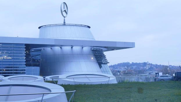 Das Mercedes-Benz Museum in Frankfurt, Deutschland, ein großes Gebäude mit einem Logo oben drauf, umgeben von Gras, Geländern, Fahrzeugen auf der Straße, Bäumen und einem klaren blauen Himmel.