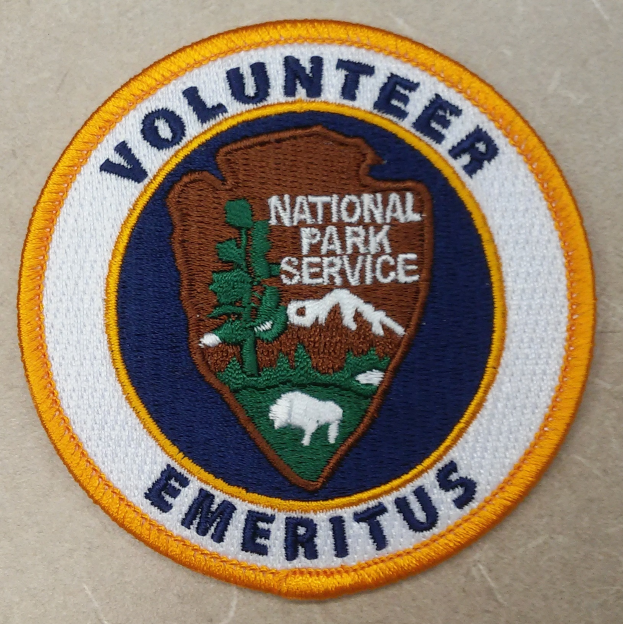 Ein runder Fleck mit einem blauen Rand, einem weißen Hintergrund und fettem schwarzem Text, der "Volunteer National Park Service Emeritus" in der Mitte eines gelben Kreises liest.