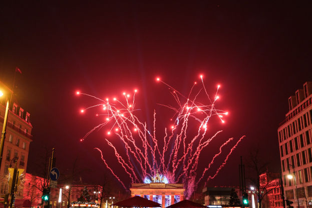 Eine Stadtstraße an Silvester in Berlin mit Gebäuden, Bäumen, Laternen, Ampeln, Schildern, Zelten, Menschen und einem Feuerwerk am Himmel.