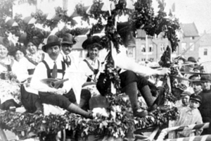 Eine Gruppe von Menschen in traditioneller bayrischer Tracht, einige mit Musikinstrumenten, fährt auf einem mit Laub geschmückten Wagen in einem Umzug, mit Gebäuden und einem klaren Himmel im Hintergrund, dargestellt in Schwarz-Weiß.