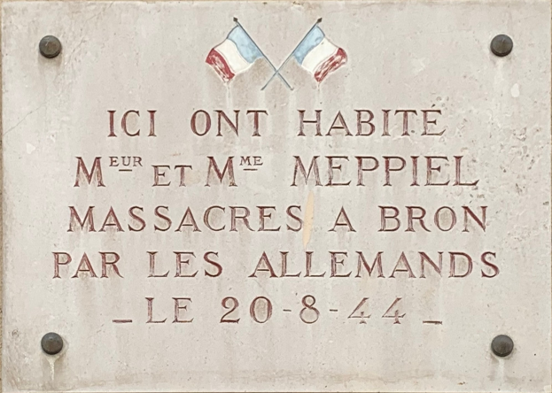 Steintafel an der Außenseite eines Gebäudes mit zwei Flaggen darüber, mit eingraviertem Text, der wahrscheinlich einen Massaker gedenkt.