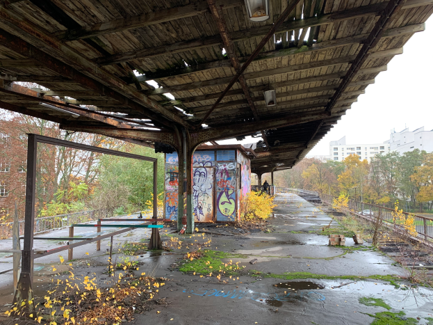 Ein verlassener Bahnhof mit Graffiti, umgeben von Bäumen, Gebäuden, Geländern, Polen, Laternen und einem Schuppen, mit trockenem Laub und Gras auf dem Boden und dem Himmel im Hintergrund.