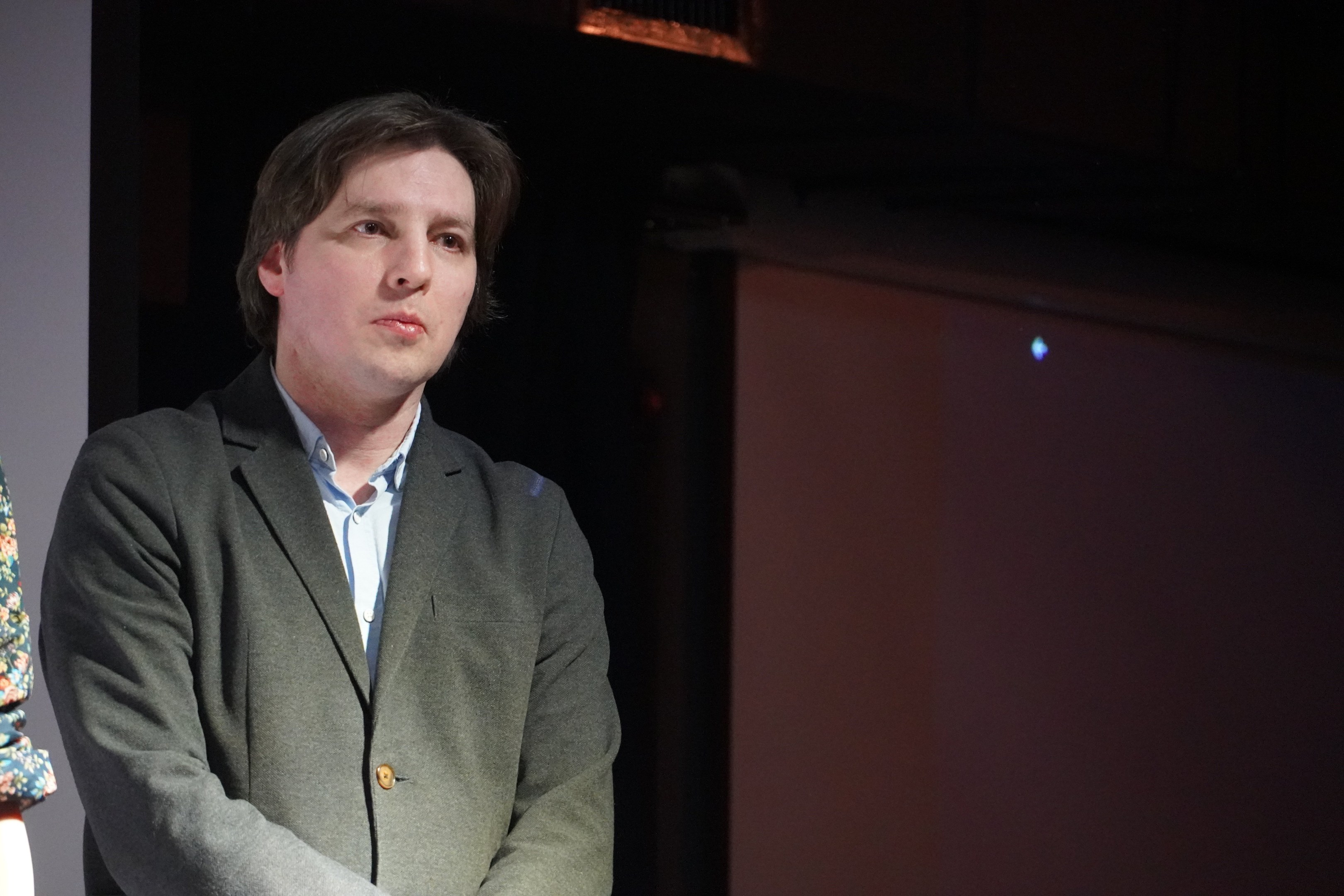 Zwei Menschen stehen nebeneinander auf einer Bühne, der Mann im Anzug und die Frau neben ihm, beide mit ernsten Gesichtern.