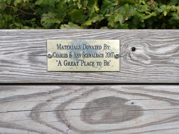 Holzbank mit einer Plakette, auf der "Materialien gespendet von Charles & Ann Schwabach 2017: Ein toller Ort" steht, vor einem Hintergrund aus üppigen grünen Pflanzen.