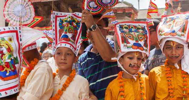 Eine Gruppe von Kindern in bunten Kostümen und Girlanden steht zusammen mit Fahnen und Gegenständen, mit Gebäuden und Pfählen im Hintergrund unter einem klaren blauen Himmel, während eines Festivals in Kathmandu.