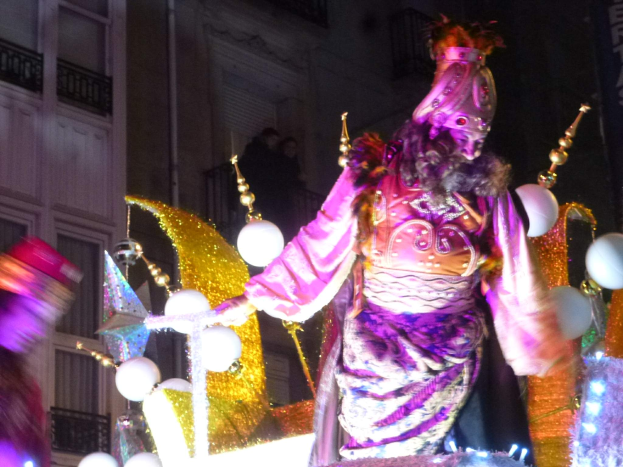 Ein Mann in einem Kostüm steht auf einem dekorierten, beleuchteten Festwagen in einem Stadtumzug, im Hintergrund ist ein Gebäude mit Fenstern und Geländern zu sehen.
